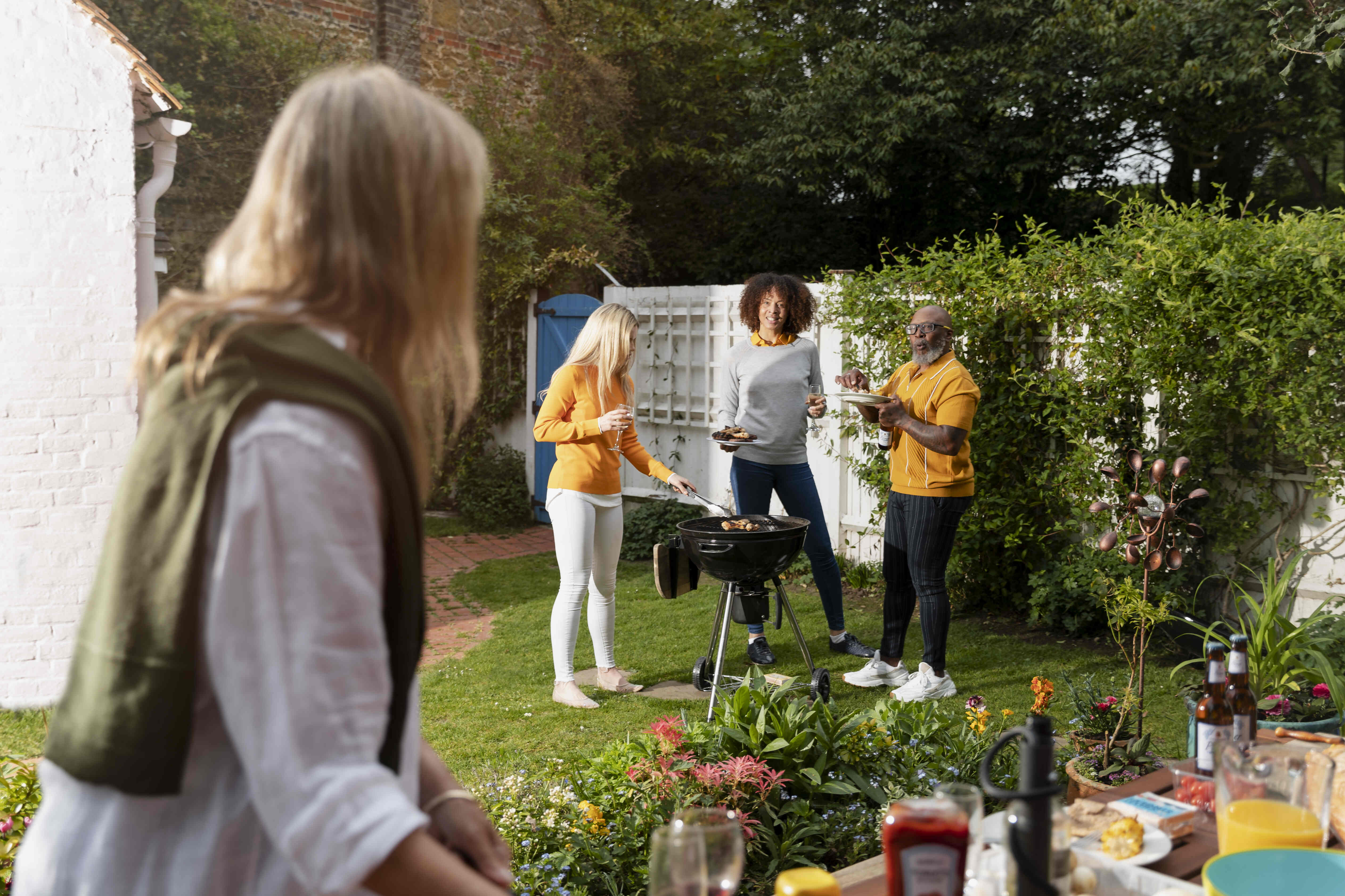 Eine Frau im Vordergrund schaut ihre Freunde an, die im Garten ein Grillfest genießen, während eine Freundin grillt und die anderen Essen und Getränke servieren.